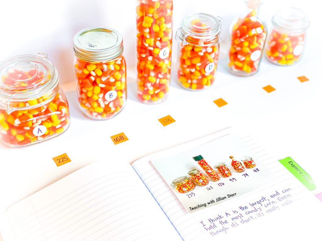 Estimation Halloween Center. 6 jars of different sizes filled with Halloween candy are in a line with a student math journal open to begin writing.