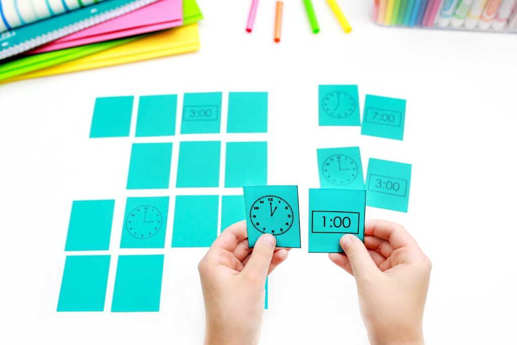 telling time to the hour game called memory match. the game is set up with cards facing down, and children's hands hold up a match between an analog clock and a digital clock reading 1:00.